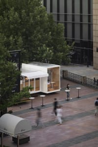 A small white building with a canopy roof surrounded by trees, with people walking around and chairs placed nearby, creating a lively atmosphere