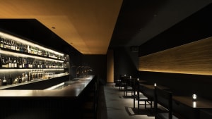 A cozy bar area with a long wooden counter, multiple bottles, and comfortable seating for patrons, set against a black wall background