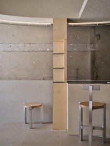 Empty modern restaurant interior with concrete walls, two stools at a counter, and a clean minimalistic atmosphere