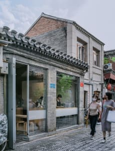 A bustling urban coffee shop with a brick facade, natural light, and people walking by on the sidewalk