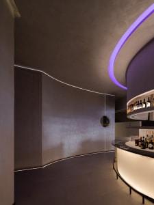 Dimly lit bar with a wooden counter and a circular clock on the wall, surrounded by bottles of alcohol