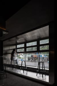A woman sits at a counter in a cafe with a glass window, surrounded by chairs and a dining table, in a calm and relaxed atmosphere