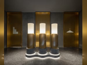 A grand hallway with three tall gold marble columns lit by side lights, set against a gray stone floor