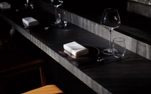 A dining table in a restaurant with a black countertop, wooden chair, wine glasses, cups, bottles, and knives