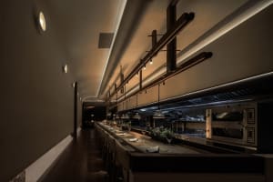 A calm and inviting restaurant dining area with a long table, chairs, and potted plants at night