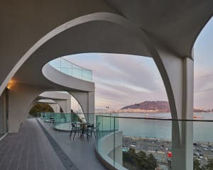 A modern building with a curved design and a large glass wall overlooking a city view, featuring a seating area with chairs and a busy urban environment outside