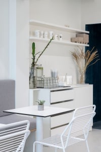 A clean and organized kitchen with a dining table and chairs, surrounded by white surfaces and a vase with a plant on the countertop
