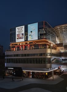 A modern Asian building at night with a large glass structure, illuminated by artificial lights, and a prominent sign on its side