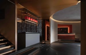 Modern hotel lobby with a circular counter area featuring a bar, sinks, and a mix of wood and tile flooring, set against a warm white and brown color scheme