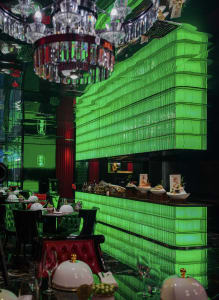 A modern restaurant interior with green walls, a bar area, and dining tables with white tablecloths and various bowls, set against a background kitchen