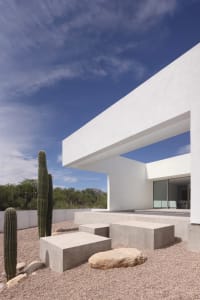 A modern house with a white exterior, featuring a large window and surrounded by cacti, suggesting a bright and open living space in a desert environment