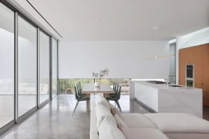 Open modern living area with white walls and floors, featuring a dining table with two chairs and a vase, a couch in the living room, and a minimalist design
