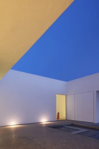 A child stands in front of a white building with a large window and two smaller windows, set in a calm and peaceful environment