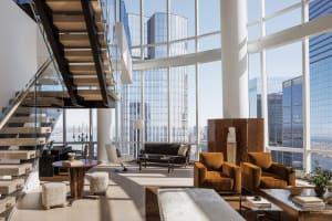 A well-lit living room in a high-rise building with hardwood floors, large windows, and comfortable seating, surrounded by potted plants and offering a view of the city skyline