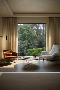 A well-lit living room with two chairs, two couches, and a dining table, surrounded by large windows and wood furniture, creating a warm and inviting atmosphere