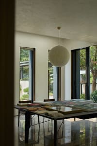 Open living area with a dining table, chairs, books, and a hanging lamp in a modern home with a predominantly white color scheme and large windows for natural light