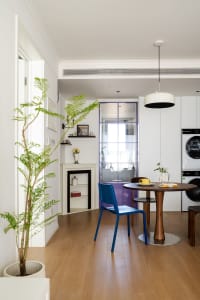 A modern kitchen and dining area with wooden floors, white walls, and a dining table surrounded by chairs and decorative items, with a potted plant in the corner