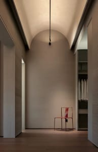 A minimalist hallway with a red chair in the center, surrounded by white walls and a single light source casting a shadow on the wall