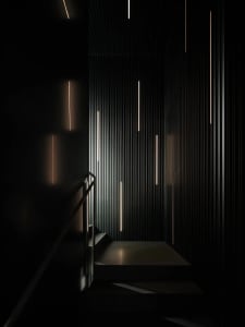 A modern stairwell with a black wall and white railing, illuminated by several lights, leading to a lower level with access to different floors