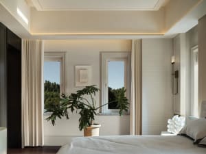 A bright bedroom with a large window, two smaller windows, and a bed extending from one end of the room to the other, with a potted plant near the foot of the bed