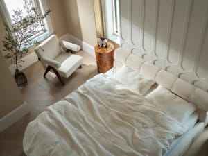 A bedroom with a large white bed on a hardwood floor, featuring two chairs, a potted plant, and a decorative vase, creating a clean and well-organized atmosphere