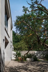 A serene outdoor space with a white building, patio, and surrounding trees, featuring a bench and two chairs for relaxation