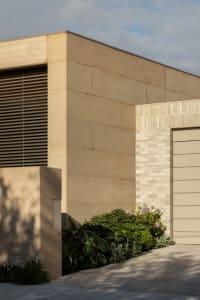 A house with a tan brick exterior, a garage door, and plants in front, set against a natural background