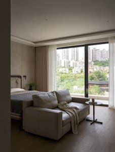 Modern living area with a white couch near a window offering a city view, wooden floors, and minimal decorations, with potted plants and a vase adding greenery and aesthetic appeal
