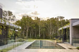 A serene outdoor pool scene with a glass-enclosed pool surrounded by trees and two houses in the background