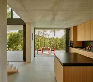 Open modern kitchen and dining space with wooden cabinets, natural light, and a dining table with cups and bowls