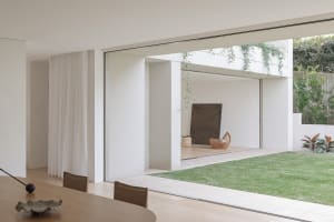 Open modern living area with glass door, two dining tables, chairs, and a couch, surrounded by white walls and minimalistic decorations