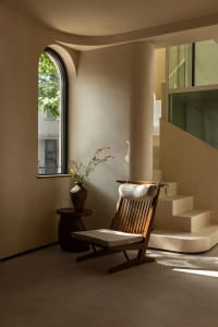 A wooden chair sits on a white carpet in front of a window, surrounded by two potted plants, creating a calm and inviting atmosphere