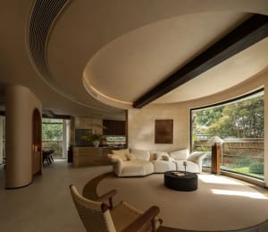 Open modern living area with a circular design, white walls, and a large window allowing natural light, featuring two couches and potted plants