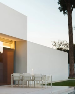 A modern outdoor patio with a dining table and white chairs, surrounded by a white building and a potted plant, creating a comfortable and inviting atmosphere