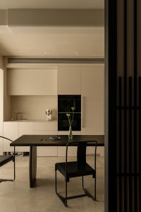 A modern kitchen with a wooden table, glass accents, and a dimly lit atmosphere, featuring a dining area with chairs, a microwave, and a vase with a flower