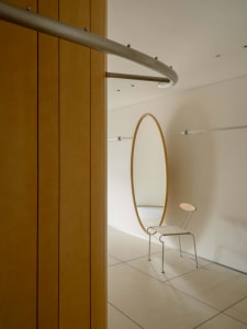 A dimly lit bathroom with a large mirror on the wall and a toilet in the background, predominantly white and minimalist in appearance