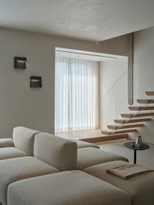 A modern living room with a couch and chair, featuring a glass staircase and large window, with natural light and a clean, minimalist appearance