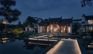 A serene and inviting Chinese garden setting with a large ornate building, a courtyard, and a pond, surrounded by stone architecture and lush greenery