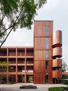 Photograph of a modern building with a brown and red color scheme, featuring a large window, spiral staircase, and brick and glass exterior, taken during the day with natural light
