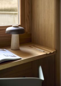 A home office with a wooden desk, chair, lamp, and book, lit by natural light from a window