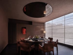 A formal dining room with a round table set for a meal, surrounded by brown chairs and a brown table, under a large window with natural light
