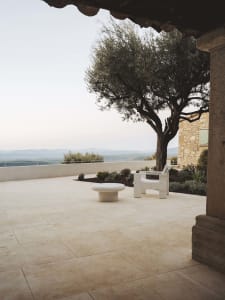 A relaxing outdoor patio with a stone wall, a white bench, and a wooden chair, surrounded by a tree and natural surroundings