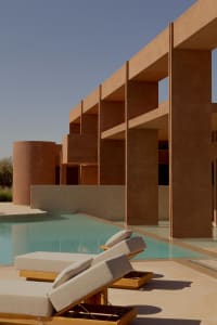 A modern house with a large window, pool, and patio area, surrounded by chairs, with natural sunlight and a warm ambiance