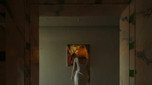 A woman in a white dress admires a painting in a room with a white wall and brown floor