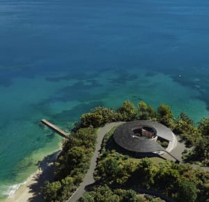 A modern house on a hillside overlooking water, surrounded by trees, with a circular structure and natural light from windows
