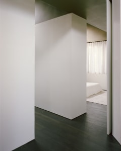 A well-lit bedroom with a white color scheme, featuring a bed near a window and a door on the right side