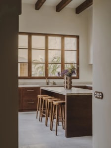 A modern kitchen with a wooden countertop, sink, refrigerator, and stools, featuring a vase with flowers and a window, with soft and warm lighting