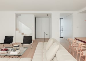 A modern living room with a white couch, black chairs, and a glass coffee table, surrounded by white walls and a staircase in the background