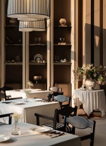 A formal dining room with a large table set with white tablecloths, wine glasses, cups, and bowls, surrounded by comfortable chairs and decorative elements like vases and potted plants, with soft and warm lighting creating a cozy ambiance