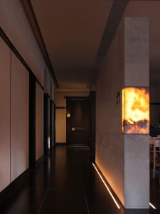 A dark hallway with a stone wall and floor, illuminated by a light fixture mounted on the wall